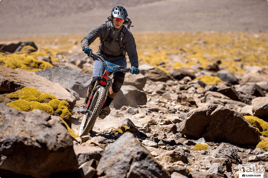 A person riding a bike uphill across rough rocky territory wearing a helmet, sunglasses,  and backpack 