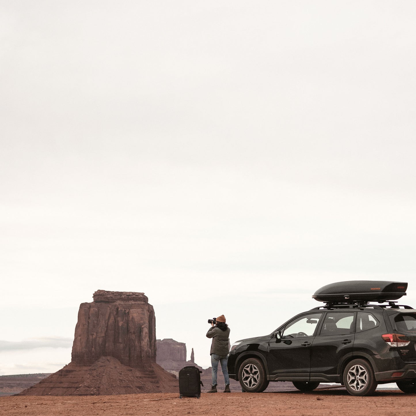 A photographer holding a camera to his face, and standing in-between a roller backpack and a black car