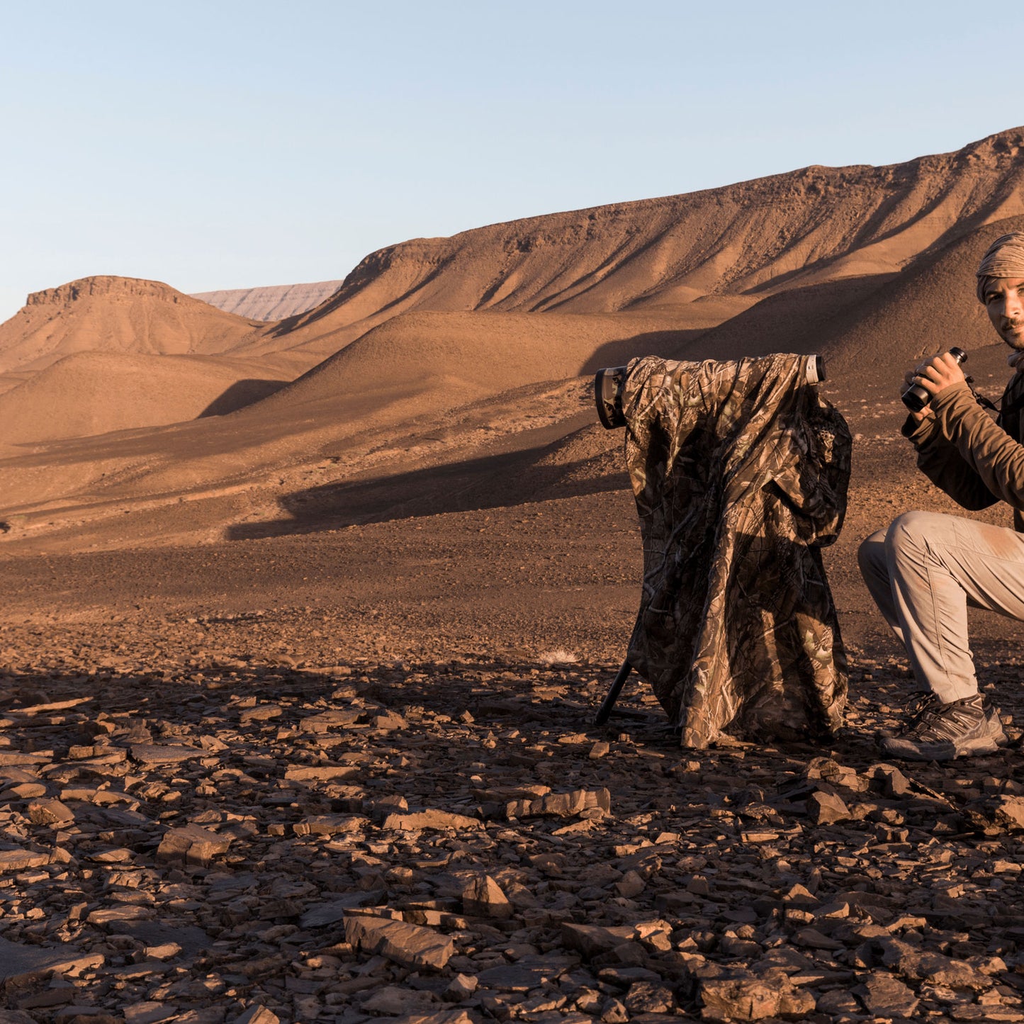 A photographer sitting holding binoculars, in front of him a camouflage tarp covering the camera with long lens 