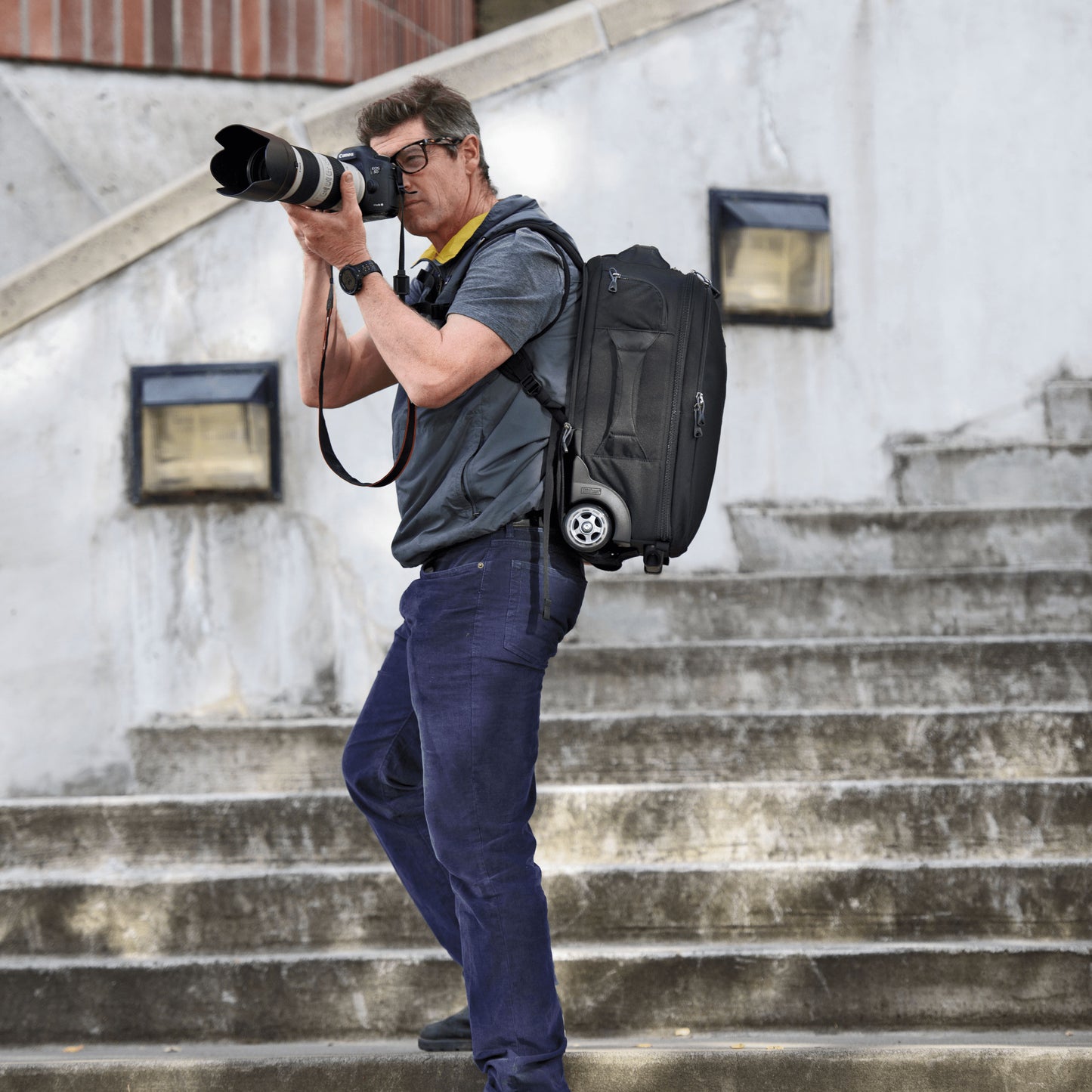 A man standing on the staircase wearing a roller backpack, holding the camera with long lens and getting ready to take a photo