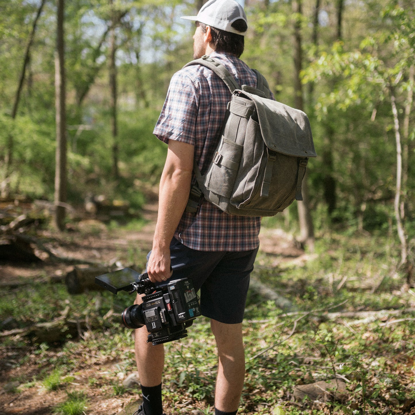 A man holding a filming equipment and wearing a backpack, gazing into the forest 