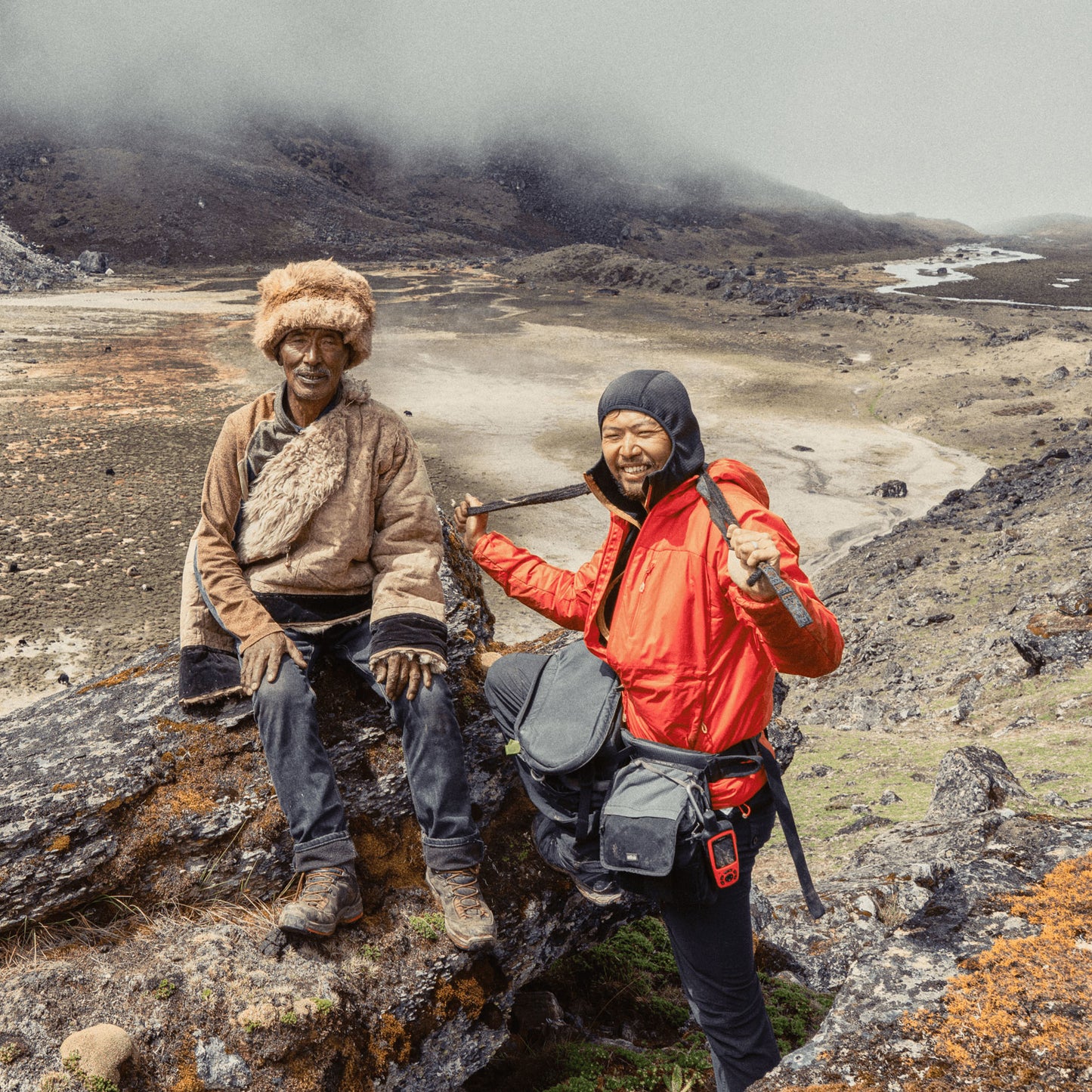 Two individuals sit on a mountainside, one in a fur hat and one in rain gear with camera equipment.