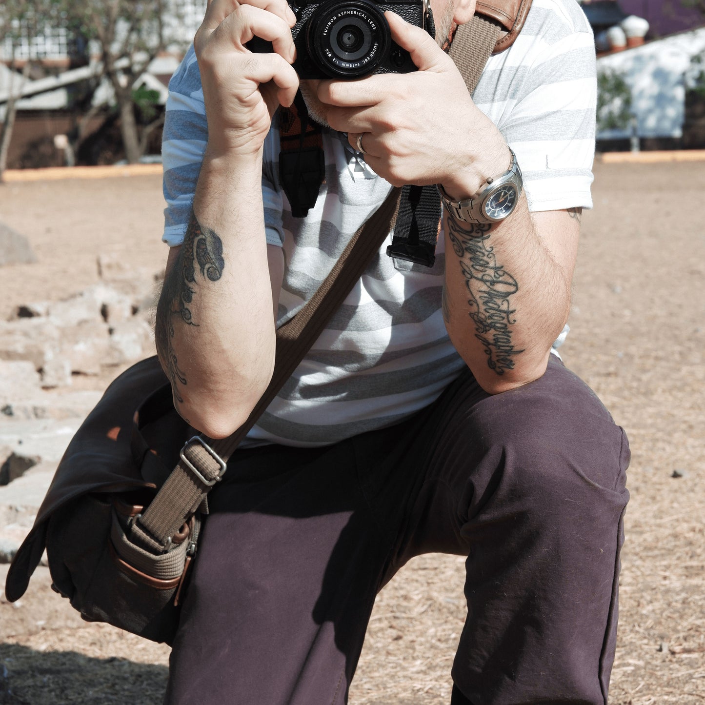 A photographer crouching down facing you, and holding a camera and taking your photo, wearing a messenger bag