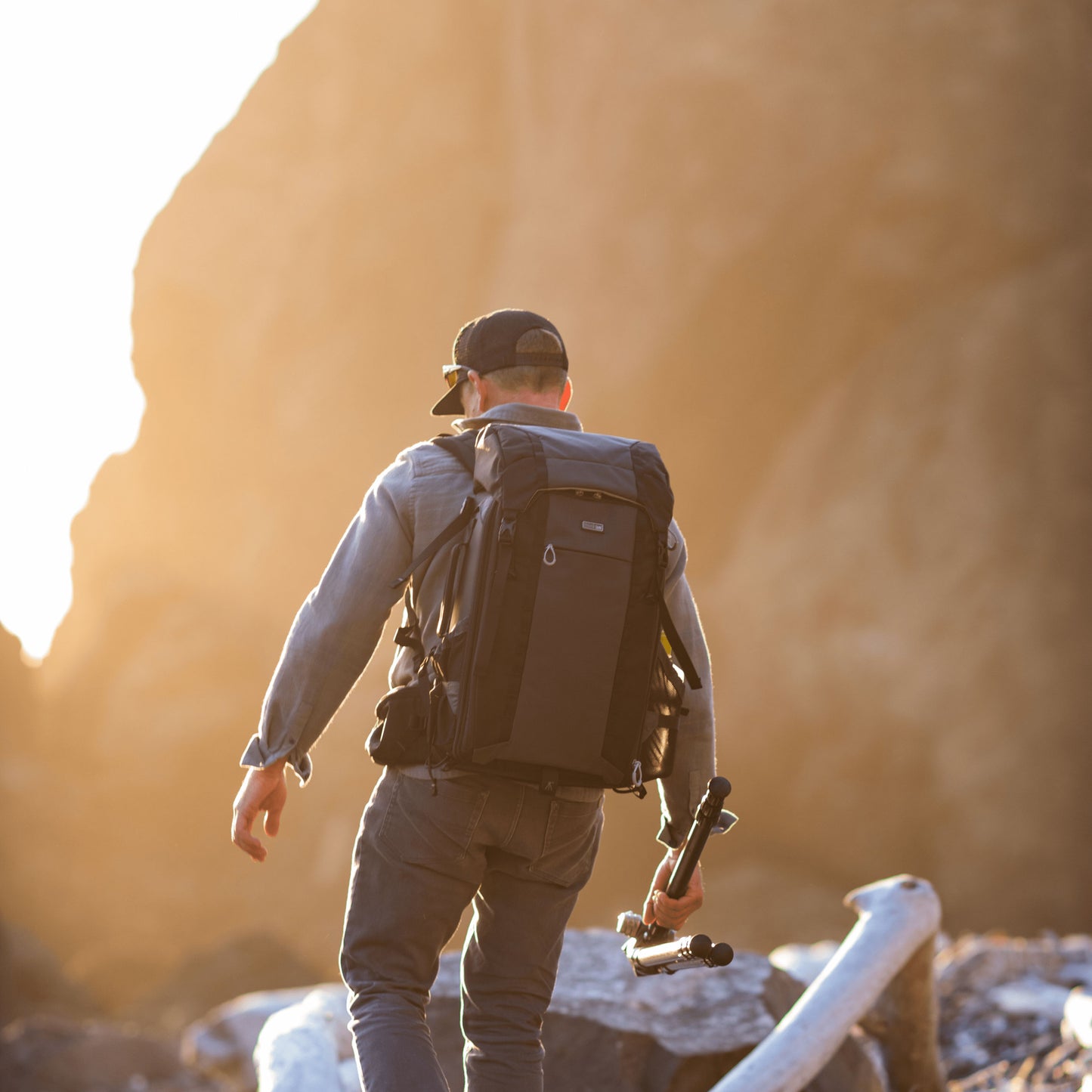 A man balancing on a log during sunrise holding a tripod and wearing a backpack