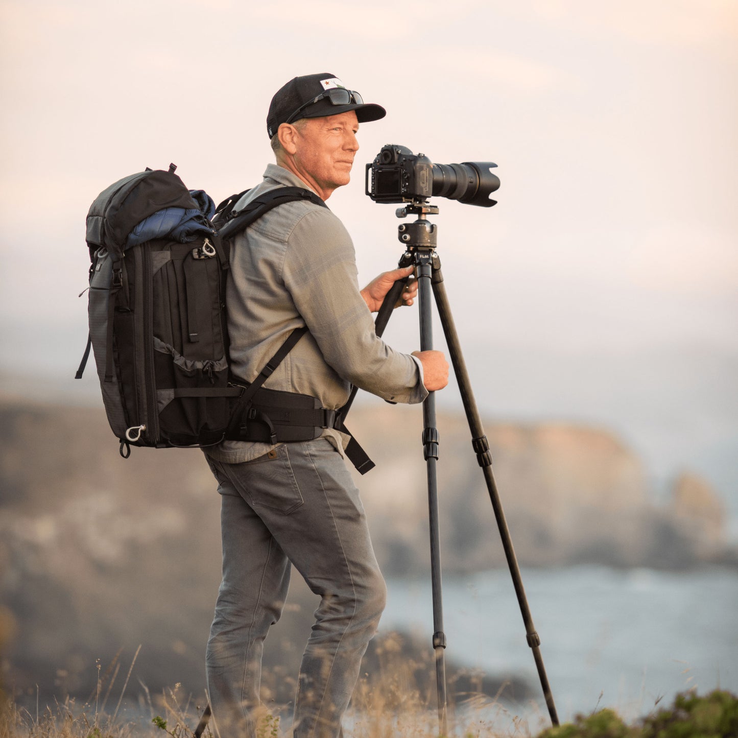 A photographer stands on a cliff wearing a backpack in front of a camera on a tripod.