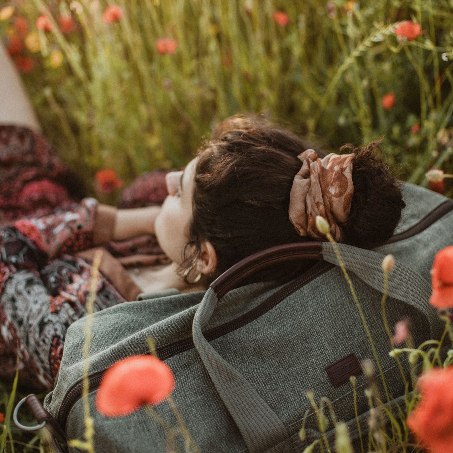 A women lying down in a field of flowers, resting her head on a duffle bag