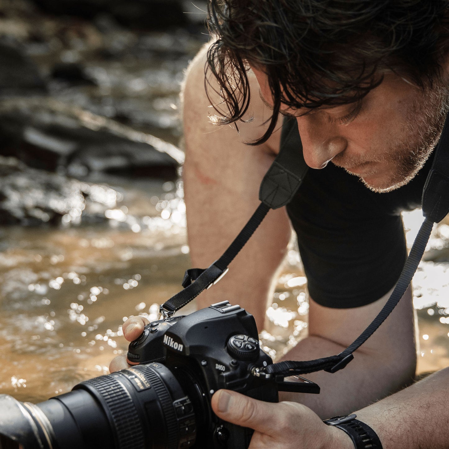 An up-close of a photographer crouching in a stream hold a camera above the water stream, positioning to take a photo 