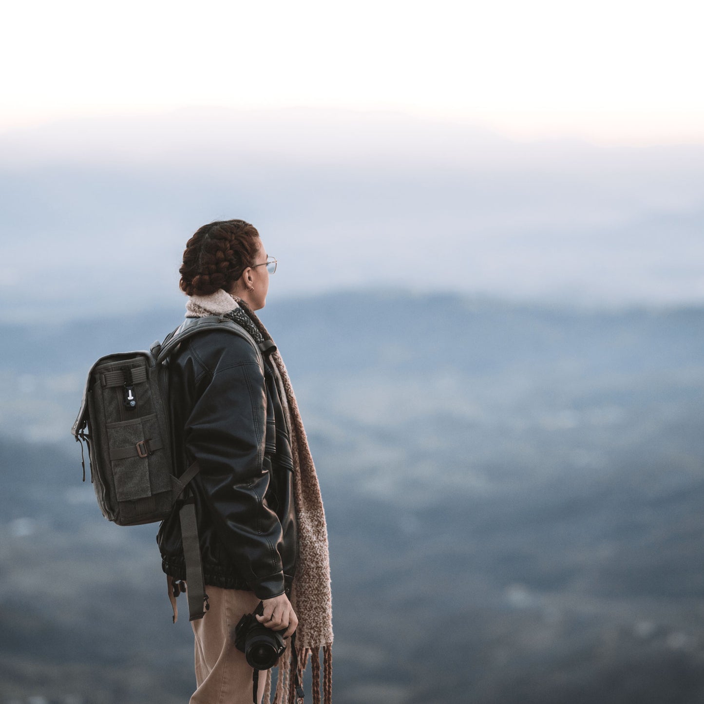 A women holding a camera equipment, and wearing a backpack and a out-of-focus mountain scenery