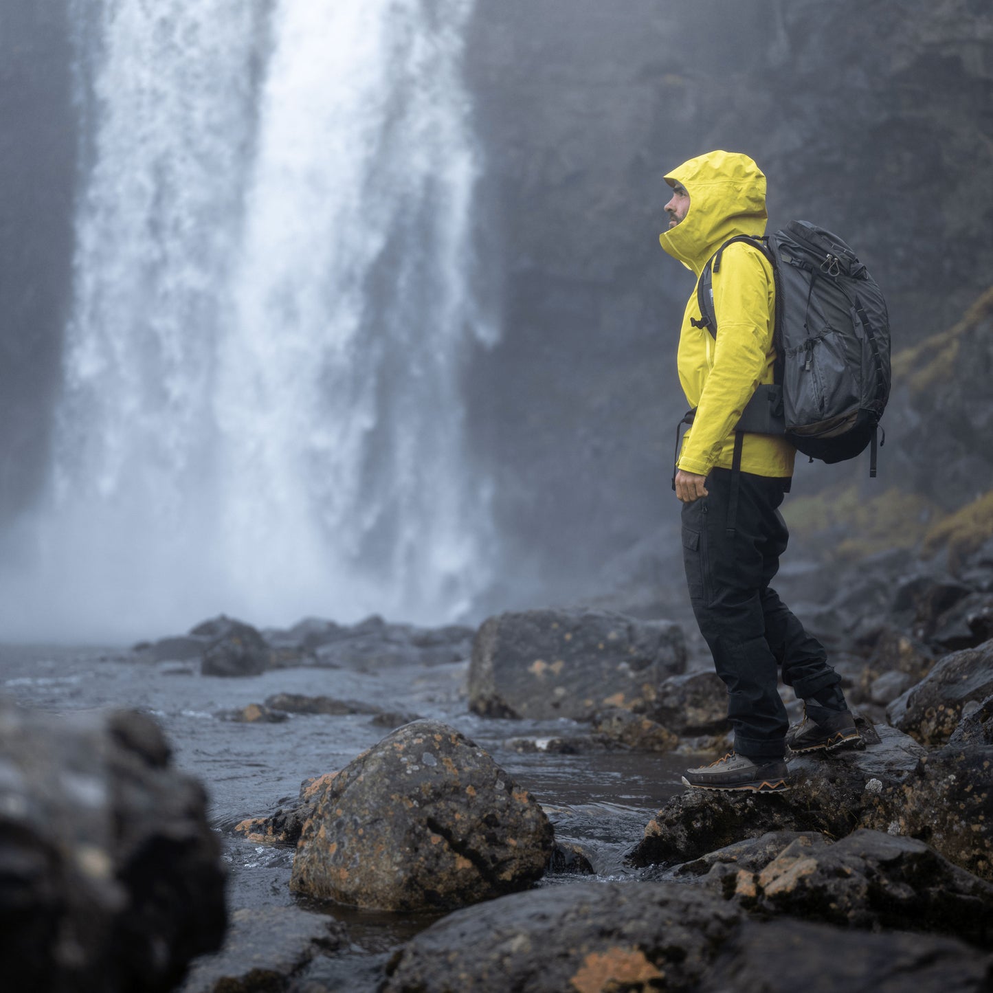 Highlands of Iceland scenery with a waterfall and a person balancing on nearby rocks wearing a raincoat and backpack