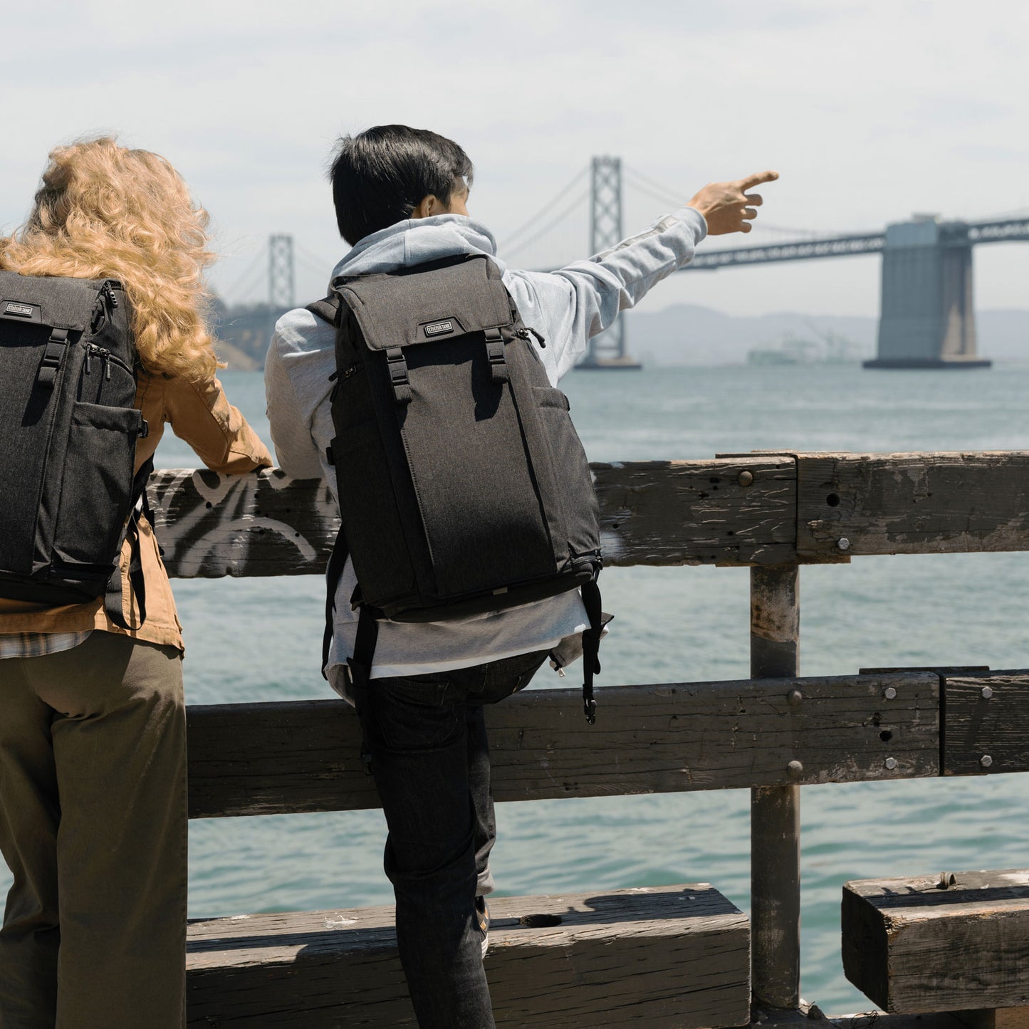 two people wearing a backpack, leaning on a fence overlooking a bridge